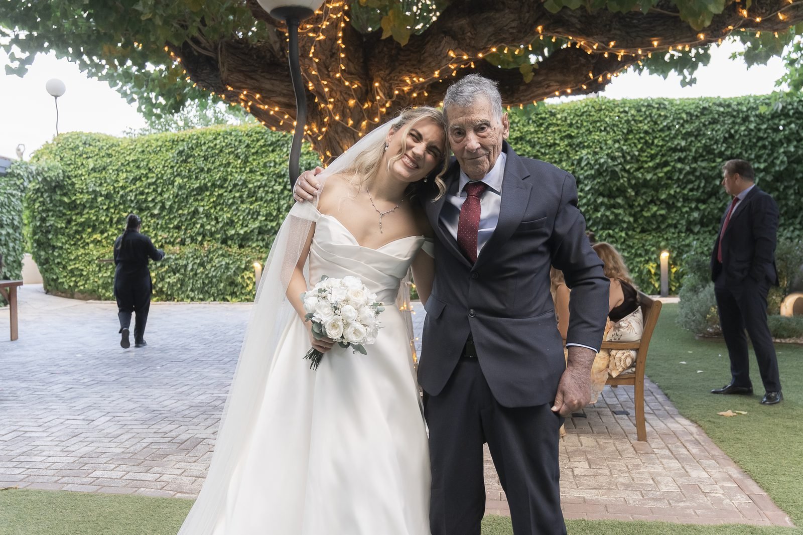 La novia abrazada a su abuelo bajo el árbol con luces del jardín de Finca Monteviejo